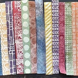 May include: A collection of colorful, patterned paper strips arranged side by side. The strips feature various designs, including floral, geometric, and abstract patterns in shades of red, blue, yellow, and green. Some strips have printed letters and numbers.