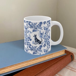 May include: White ceramic mug with a blue and white floral pattern and a black silhouette of a dog sitting in the center. The mug has a handle.