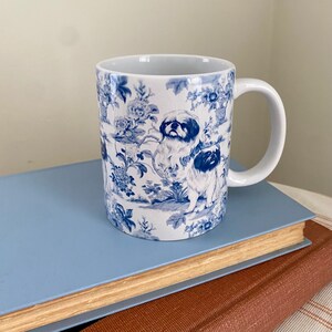 May include: A white ceramic mug with a blue and white floral pattern featuring a dog breed. The mug has a handle and is sitting on a blue book.