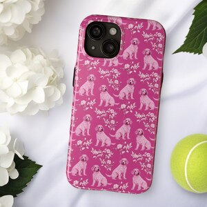 May include: A pink phone case with a white floral pattern and a repeating image of a white dog. The phone case has a cutout for the camera lens.