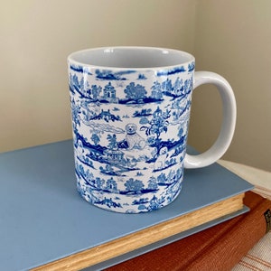 May include: A white ceramic mug with a blue and white pattern of dogs and houses. The mug has a handle and is sitting on a blue and brown surface.