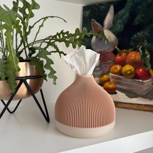 May include: A peach and cream-colored tissue box with a ribbed design, holding white tissues. A green plant in a copper pot sits on a black metal stand to the left. A painting of a rabbit and fruit is in the background.