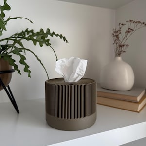 May include: An olive green tissue box with a ribbed design. The cylindrical container has a solid base and a top with a central opening, dispensing white tissues. The box sits on a white shelf, with a plant and vase in the background.