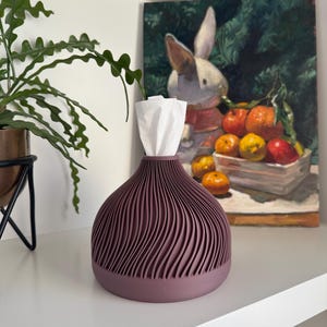May include: A mauve tissue box with a ridged, wave-like design. The tissue box is on a white shelf, with a painting of a rabbit and fruit in the background. A green plant in a black metal pot is to the left.