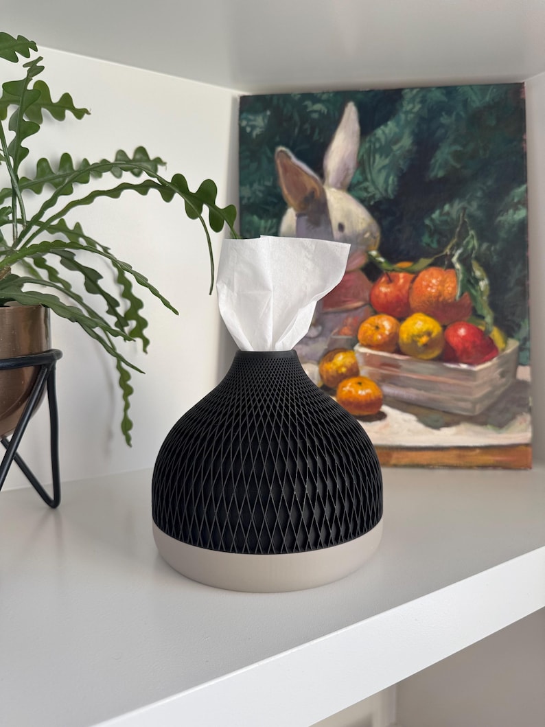May include: A black and beige tissue box with a geometric design. The tissue box is on a white shelf, with a painting of a rabbit and fruit in the background. The tissue box has a black, diamond-patterned top and a beige base.