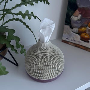 May include: A decorative tissue box with a geometric pattern in beige and a dark purple base. White tissues are visible at the top. The tissue holder is on a white shelf, with a green plant and a painting in the background.