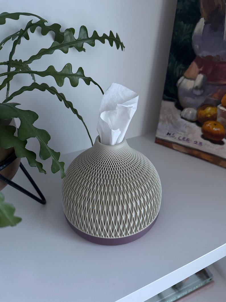 May include: A decorative tissue box with a geometric pattern in beige and a dark purple base. White tissues are visible at the top. The tissue holder is on a white shelf, with a green plant and a painting in the background.