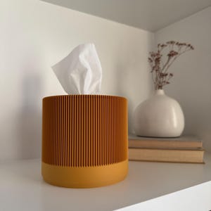 May include: A cylindrical tissue box with a ribbed, orange exterior and a mustard-yellow base. White tissues are pulled from the top. The tissue holder sits on a white shelf, next to a white vase and books.