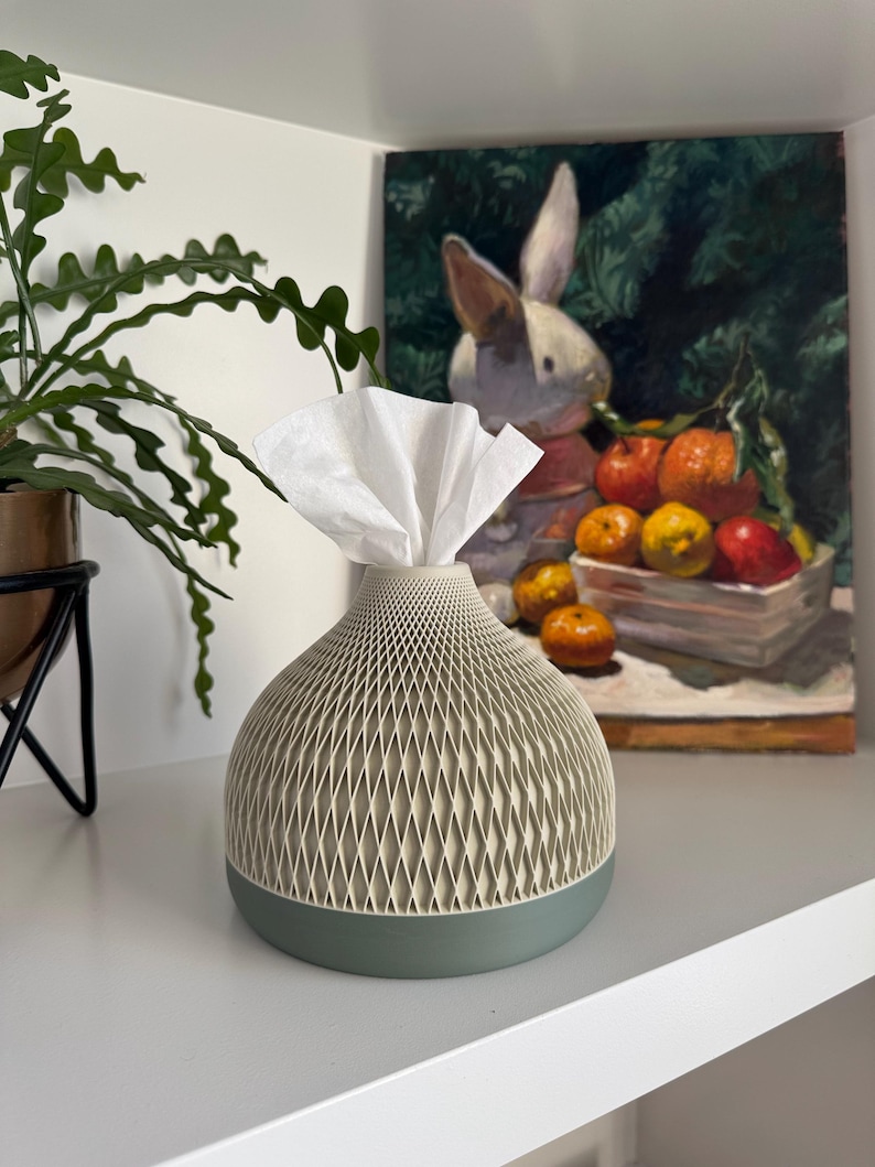 May include: A decorative tissue box with a geometric pattern in cream and a sage green base. White tissues are pulled from the top. The tissue holder is on a white shelf, with a painting of a rabbit and fruit in the background.