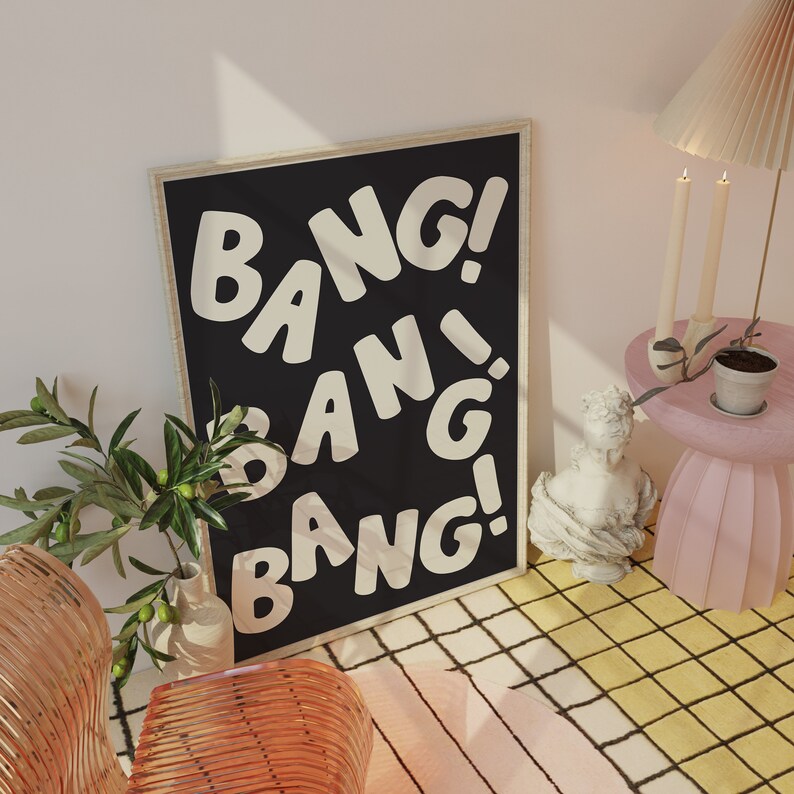 Pu&ograve; includere: Un poster incorniciato nero con testo bianco che recita "Bang! Bang! Bang!" Il poster &egrave; appeso a una parete bianca con una pianta in un vaso e un tavolino rosa con una scultura sopra.