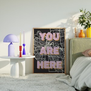 May include: A black and white city map print with the words "YOU ARE HERE" in pink lettering. The print is framed and hanging on a wall above a bed with a green headboard. There are candles and a lamp on a white table in front of the print.