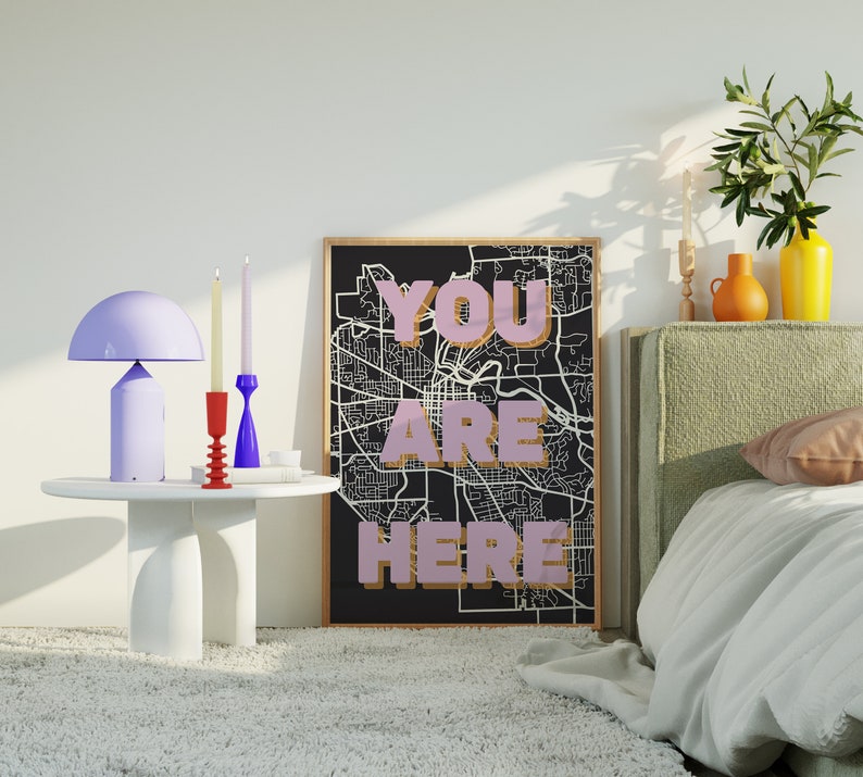 May include: A black and white city map print with the words "YOU ARE HERE" in pink lettering. The print is framed and hanging on a wall above a bed with a green headboard. There are candles and a lamp on a white table in front of the print.