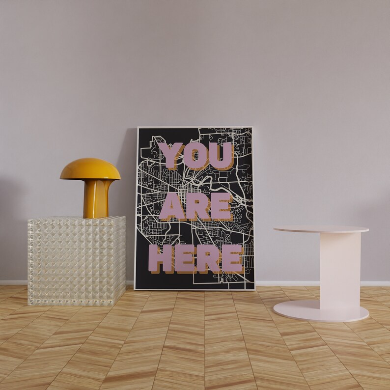 May include: A black and white map print with the words "YOU ARE HERE" in pink letters. The print is framed and hanging on a light purple wall. A yellow lamp and a white side table are in the foreground.