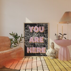 May include: A black and white map print with the words "YOU ARE HERE" in pink letters. The print is framed and hanging on a wall in a room with a pink and yellow rug, a pink side table, and a pink chair.