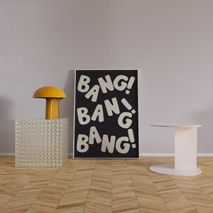 Pu&ograve; includere: Un poster incorniciato nero con testo bianco che recita "BANG! BANG! BANG!" Il poster &egrave; appeso a una parete di colore lilla chiaro. In primo piano, una lampada gialla e un tavolino bianco.
