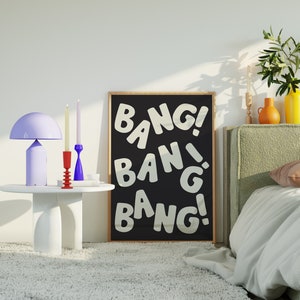 Pu&ograve; includere: Un poster incorniciato nero con testo bianco che recita "Bang! Bang! Bang!" Il poster &egrave; appeso a una parete bianca sopra un letto con una testiera verde. Ci sono due candelabri e una lampada su un tavolino bianco.