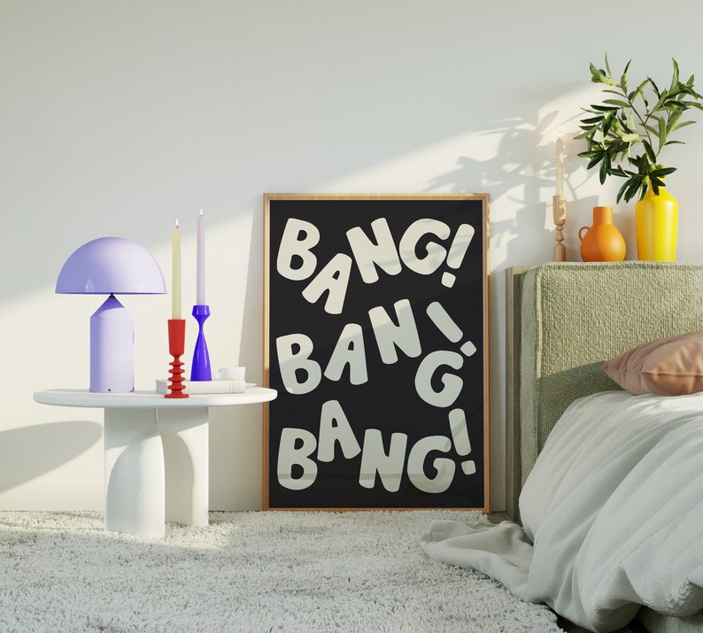 Pu&ograve; includere: Un poster incorniciato nero con testo bianco che recita "Bang! Bang! Bang!" Il poster &egrave; appeso a una parete bianca sopra un letto con una testiera verde. Ci sono due candelabri e una lampada su un tavolino bianco.