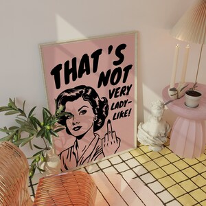 K&ouml;nnte beinhalten: Ein rosa und wei&szlig;es Poster mit einer Schwarzwei&szlig;illustration einer Frau, die den Mittelfinger zeigt. Der Text auf dem Poster lautet "That's not very lady-like!"