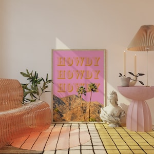 May include: A pink framed poster with the word "HOWDY" repeated three times in a retro font. The poster features a desert landscape with palm trees and mountains. The poster is hanging on a pink wall.