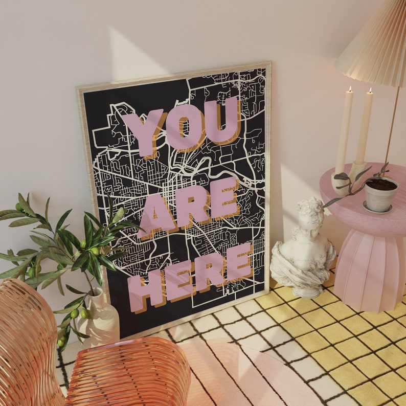 May include: A black and white map print with the words "YOU ARE HERE" in pink letters with a gold outline. The print is framed and hanging on a white wall.