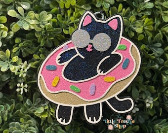 May include: A black cat wearing sunglasses floats in a pink donut with colorful sprinkles. The cat has glittery accents and is surrounded by a green leafy background. The product is from "the little Freshe Shop".