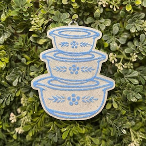 May include: A light-colored ornament shaped like a stack of three vintage mixing bowls with blue trim and floral and leaf designs. The ornament has a silver loop for hanging and is set against a backdrop of green foliage.