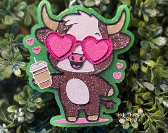 May include: A glittery brown cow-shaped charm wearing pink heart-shaped sunglasses and holding a coffee drink. The cow has a white face and a green outline. Pink hearts surround the cow. The charm has the text "Little Freshe Shop".