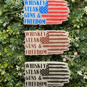 May include: Three flag-shaped ornaments with the text "WHISKEY STEAK GUNS & FREEDOM." The top ornament has a blue and red American flag design. The middle ornament has a brown flag design. The bottom ornament has a black and white flag design.
