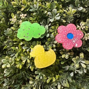 May include: Three colorful, decorative resin shapes: a green cloud, a pink flower with a blue center, and a yellow duck. The shapes are sprinkled with glitter and rest on a bed of green foliage.