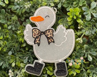 May include: A glittery gray duck-shaped ornament with an orange beak and a leopard print bow tie. The duck is wearing black boots with glitter accents. The ornament is set against a backdrop of green foliage.