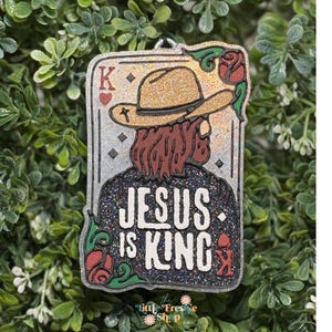May include: A rectangular pendant with a glittery background and the words "JESUS IS KING". The pendant features an illustration of a figure wearing a cowboy hat and a playing card design with a red rose.