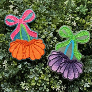 May include: Two glittery seashell-shaped ornaments. One has a pink bow and orange shell, the other has a green bow and purple shell. Both have blue and green accents. The ornaments are set against a backdrop of green foliage.