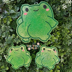 May include: Three green frog-shaped air fresheners with a glittery finish, black eyes, and pink cheeks. The largest frog is positioned above two smaller identical frogs. The text "The Little Freshie Shop" is printed in the center between the frogs.