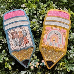 May include: Two pencil-shaped ornaments with glitter accents. One is blue with the text "Teach Her", the other is gold with a rainbow and the word "Teacher". Both have pink erasers and silver accents.