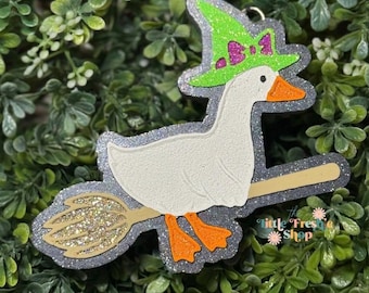 May include: A whimsical, glittery charm featuring a white goose riding a broom, wearing a green witch hat with a purple bow. The charm has orange feet and beak, and a silver glitter outline. The broom is gold glitter. The charm is set against a green leafy background.