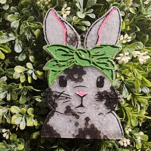 May include: A bunny-shaped ornament with a green bow and pink-lined ears. The bunny has a gray and black speckled pattern and a pink nose. The ornament is set against a backdrop of green foliage and white flowers.