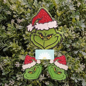 May include: Glittery green Grinch Christmas ornaments with red Santa hats and white trim. The Grinch's face has a grumpy expression with yellow eyes. A small card with text is in the center. Festive holiday decor.