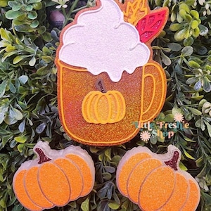 May include: Glittery orange pumpkin spice latte and pumpkin-shaped ornaments. The latte features whipped cream and a pumpkin design. The pumpkins are orange with glitter and brown stems. The items are set against a green leafy background.