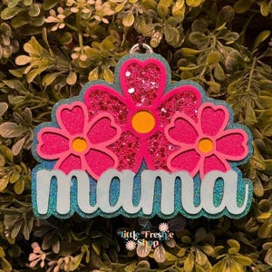 May include: A floral-shaped keychain with the word "mama" in white script. The keychain features three pink flowers with yellow centers, set against a glittery pink background. The base is a light blue with a glittery effect.