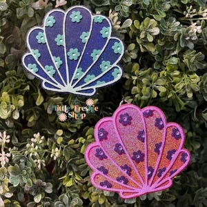 May include: Two seashell-shaped keychains. One is dark blue with light blue flower accents, the other is pink with purple flower accents. The keychains have glitter accents and are set against a background of green foliage. The text "the little Freshe Shop" is visible.