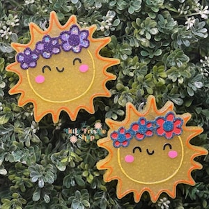 May include: Two smiling sun-shaped ornaments with orange rays and yellow centers. Each sun has a floral crown and pink cheeks. The background is a bed of green foliage. The text "the little Freshe Shop" is visible.