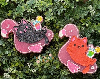 May include: Two resin flamingo float ornaments, one with a black glitter cat and the other with an orange cat, each holding a cocktail. The flamingos are pink with glitter accents, set against a green leafy background.