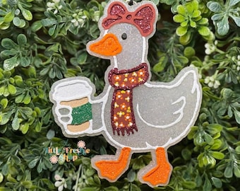May include: A whimsical, glittery goose ornament with a red bow, scarf, and orange feet and beak. The goose is holding a coffee cup with a green sleeve. The ornament has a white outline and is set against a green leafy background.