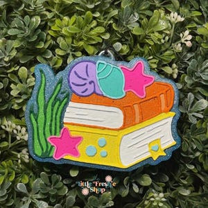 May include: A colorful, glittery ornament featuring a stack of books with a seashell and starfish design. The ornament has a blue border and includes green seaweed and the text "little Freshie Shop".