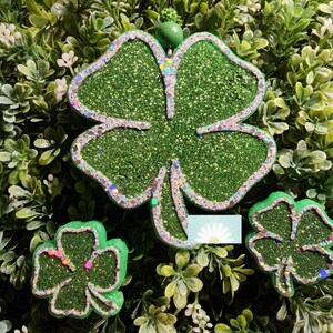 May include: A set of three green glitter shamrock decorations. The largest shamrock has a silver glitter outline and a green bead at the top. The smaller shamrocks also have glitter accents. The background is a bed of green foliage.