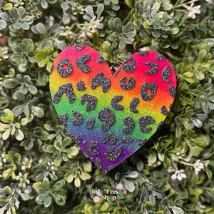 May include: A heart-shaped keychain with a rainbow gradient background and black glitter leopard print spots. The keychain is set against a backdrop of green foliage. The text "Little Fresh Shop" is visible at the bottom.