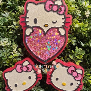 May include: Three Hello Kitty-themed resin decorations. The largest features a heart filled with pink glitter. The other two are smaller Hello Kitty faces. All have red outlines, white faces, and pink bows. The text "little Fresh Shop" is visible.