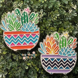 May include: Two colorful cactus-themed decorations. Each features a potted succulent with a zig-zag pattern. The plants have green, orange, and blue leaves. The pots are orange and purple with glitter accents. The text "little tresie shop" is visible.