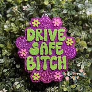 May include: A purple glittery keychain with the text "DRIVE SAFE BITCH" in green glitter. The keychain is shaped like a flower and has smiley faces and flower accents. The words "Little Freshie Shop" are also visible.