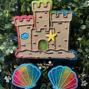 May include: A sandcastle-shaped ornament with a yellow starfish and a blue seashell. The sandcastle has a textured, tan surface with colorful trim. Two seashell-shaped ornaments with rainbow gradients are also visible.